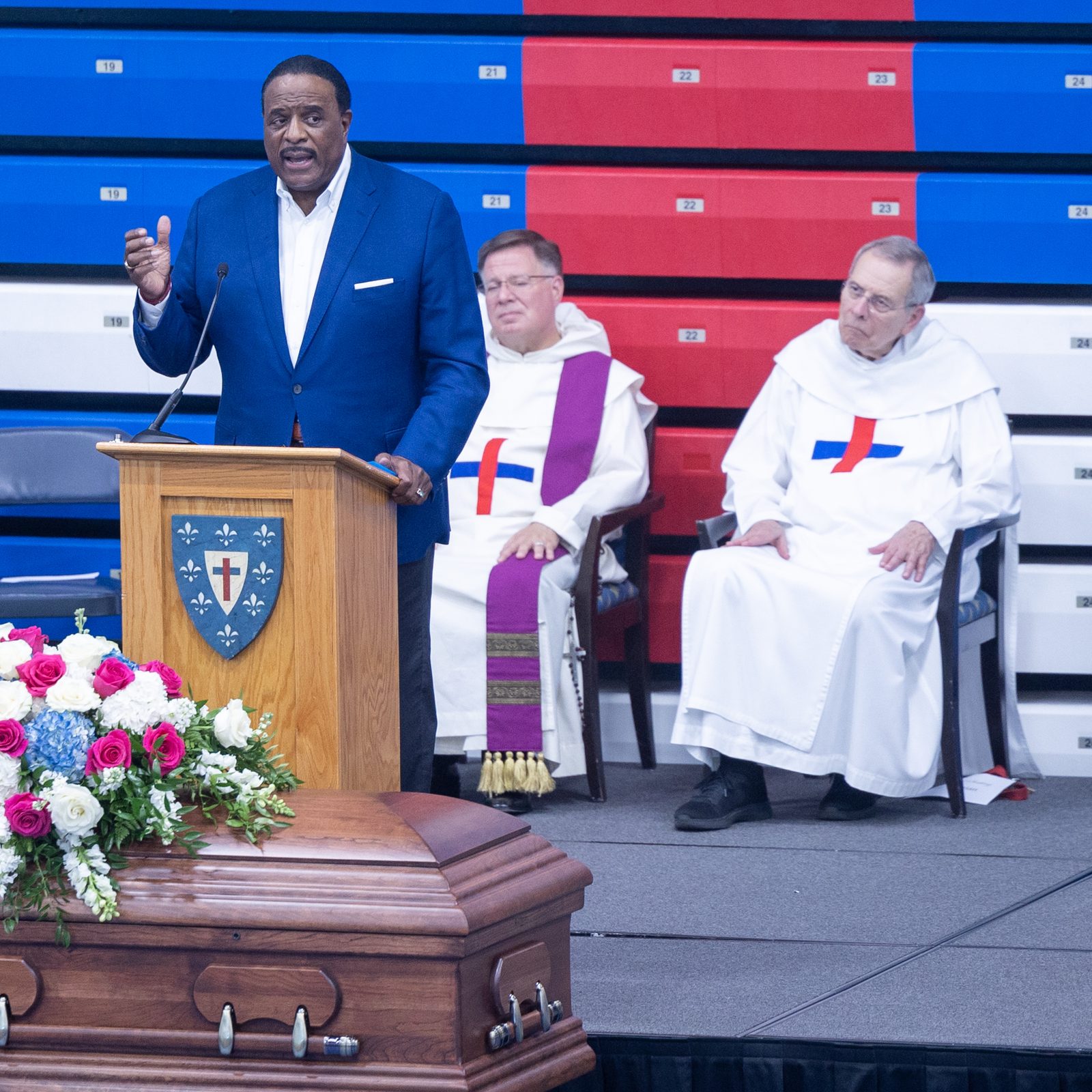 James Brown, the longtime anchor for “The NFL Today” on CBS, speaks at a vigil service on July 21, 2025 at the Lt. (SEAL) Brendan Looney Convocation Center at DeMatha Catholic High School in Hyattsville for fellow DeMatha graduate Tim Strachan, who died on July 8 at the age of 49 after suffering from cancer. At left is Johnny Holliday, who partnered with Strachan on radio broadcasts for Maryland Terrapins football games. At center is Trinitarian Father Albert Anuszewski, the president of DeMatha Catholic High School, and at right is Trinitarian Father Damian Anuszewski, a longtime DeMatha teacher who led the closing prayers at the vigil. (Catholic Standard photo by Mihoko Owada)