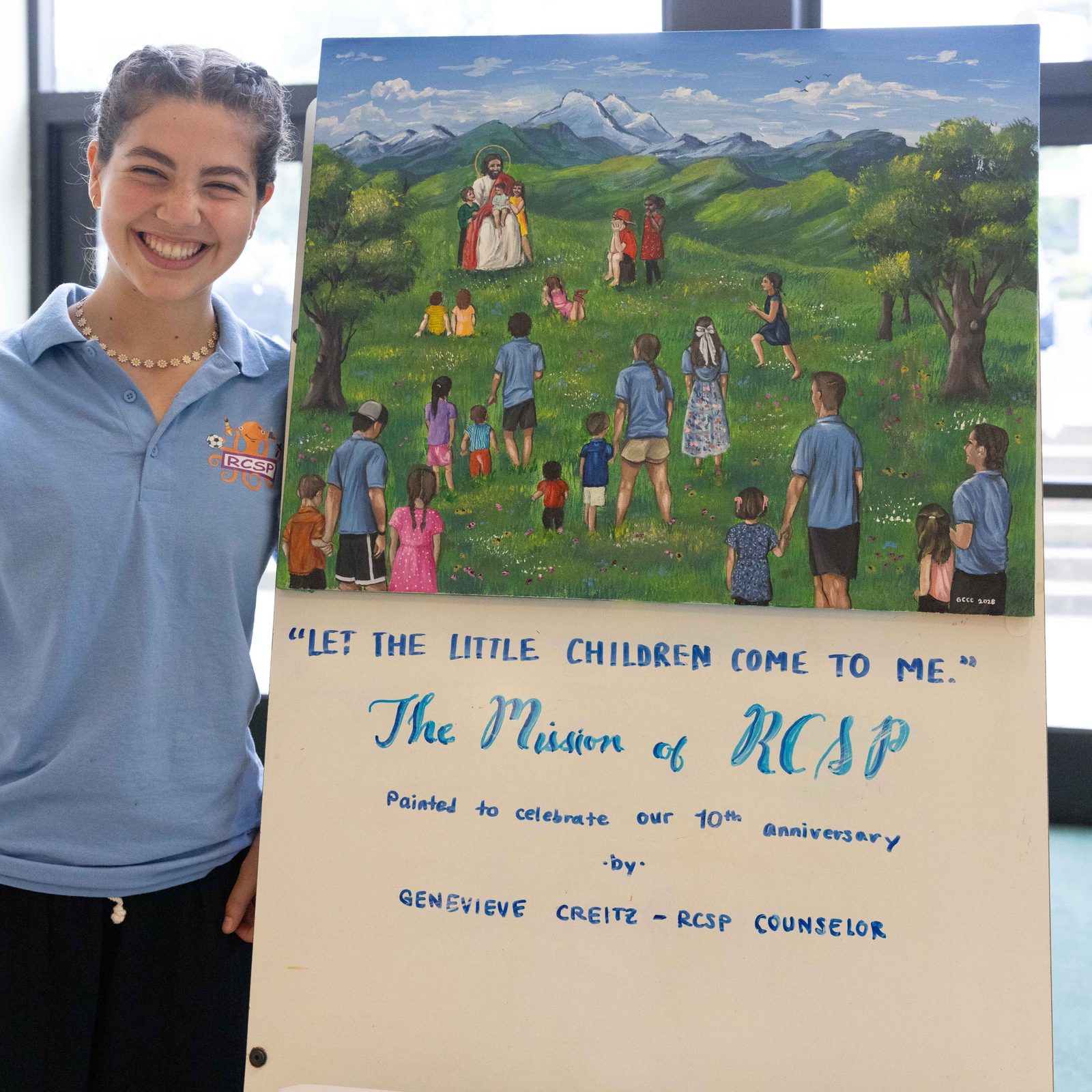 Genevieve Creitz, a counselor at the Rockville Catholic Summer Program at St. Patrick’s Parish in Rockville, Maryland, stands next to a painting titled “Let the Little Children Come to Me,” that she painted to mark the camp’s 10th anniversary in 2026. The painting shows campers and counselors at the camp walking to Jesus. Creitz, who is 17, is interested in studying sacred art and iconography in college. (Catholic Standard photos by Andrew Biraj)