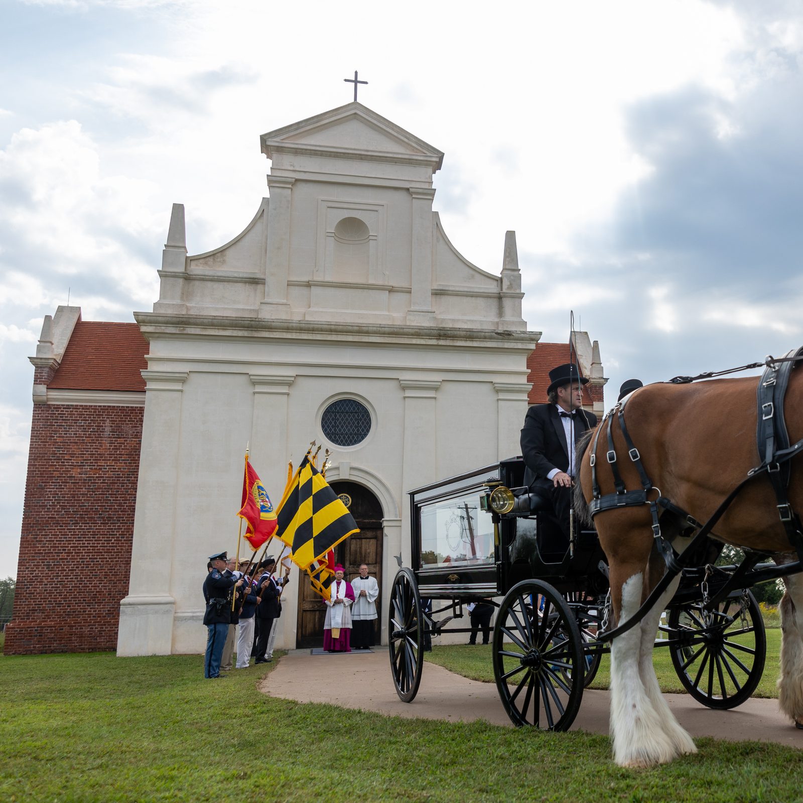 The hearse bearing the remains of eight young children from colonial-era Maryland arrives at the rebuilt 1667 Brick Chapel in Historic St. Mary’s City on Sept. 20, 2025 after being pulled by two Clydesdale horses in a procession from the reconstructed State House of 1676. Baltimore Archbishop William E. Lori, who presided at a reinterment ceremony at the Brick Chapel, can be seen in the background waiting at the chapel’s entrance. The Brick Chapel stands as a landmark of faith that symbolizes Maryland’s status as a birthplace of religious freedom in the United States. (Catholic Standard photo by Mihoko Owada)