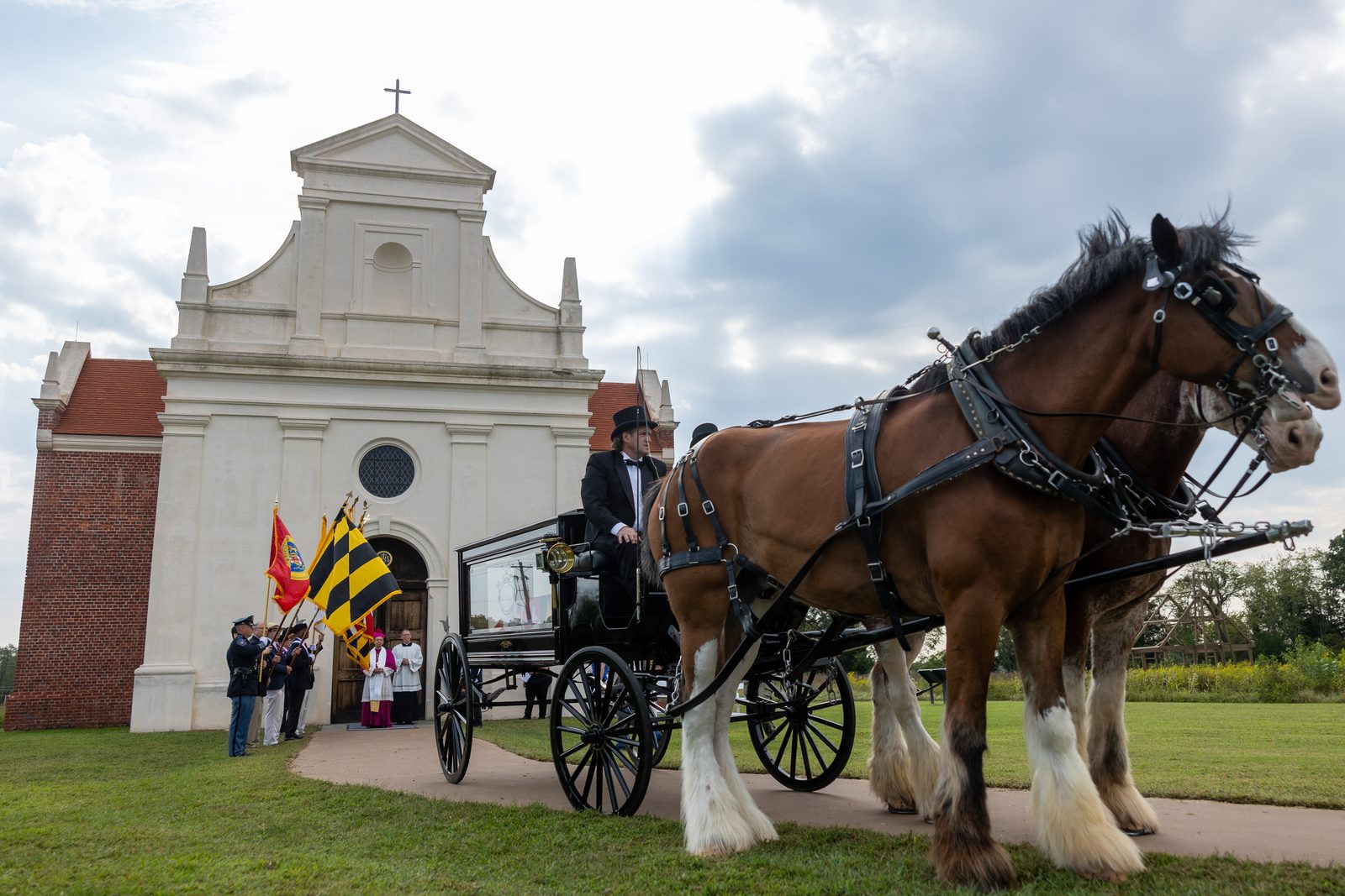 The hearse bearing the remains of eight young children from colonial-era Maryland arrives at the rebuilt 1667 Brick Chapel in Historic St. Mary’s City on Sept. 20, 2025 after being pulled by two Clydesdale horses in a procession from the reconstructed State House of 1676. Baltimore Archbishop William E. Lori, who presided at a reinterment ceremony at the Brick Chapel, can be seen in the background waiting at the chapel’s entrance. The Brick Chapel stands as a landmark of faith that symbolizes Maryland’s status as a birthplace of religious freedom in the United States. (Catholic Standard photo by Mihoko Owada)