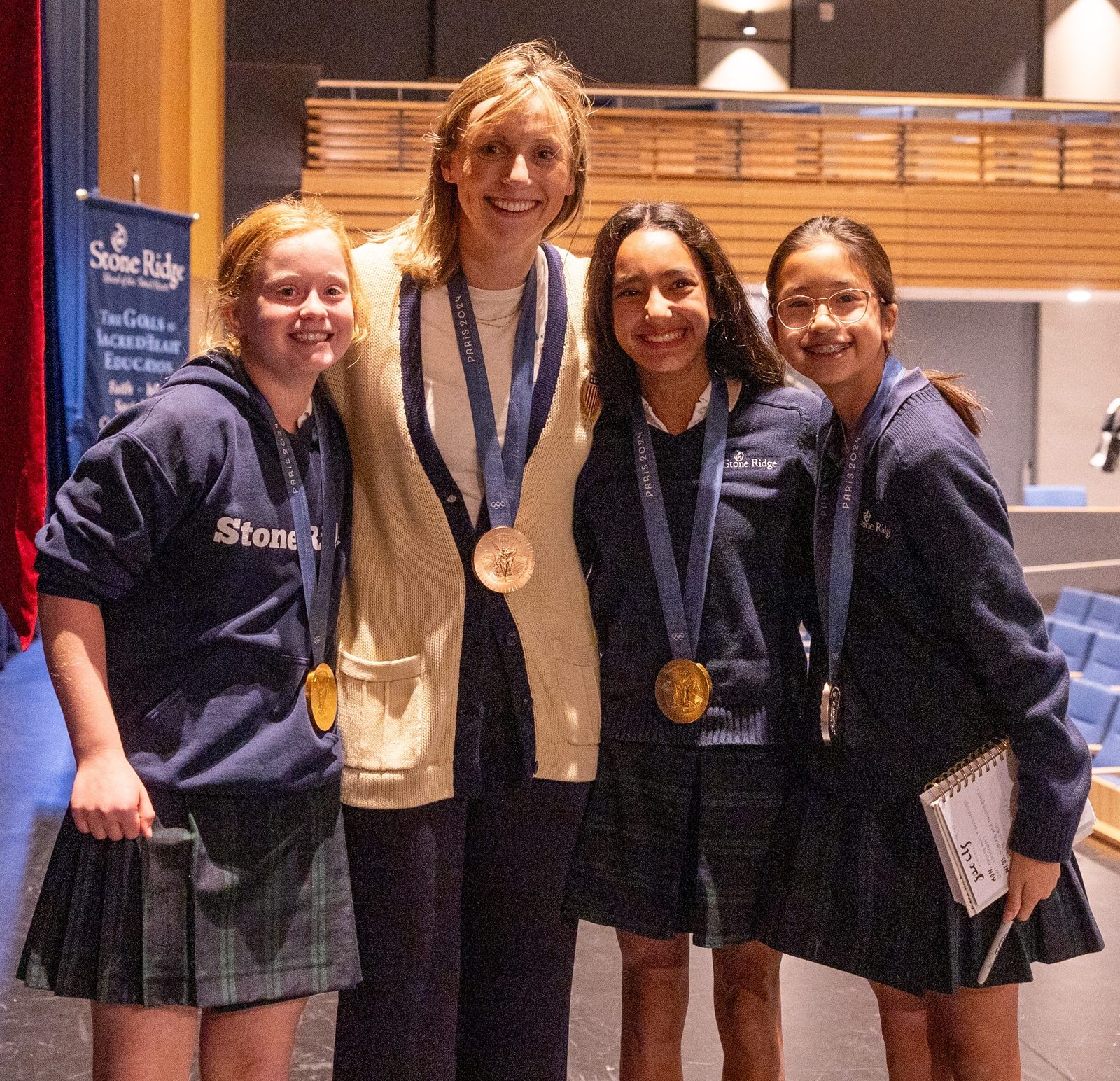 After Olympic champion swimmer Katie Ledecky spoke to Middle School students during her visit to her alma mater, Stone Ridge School of the Sacred Heart in Bethesda, Maryland, on Oct. 22, 2024, she let some students wear her medals from the recent Summer Olympic Games in Paris. From left to right are student Hope Murphy, Ledecky, and students Darya Tamiz and Eliana Liu. At the Paris Olympics, Ledecky won two gold medals, a silver medal and a bronze medal.  (Catholic Standard photo by Mihoko Owada)