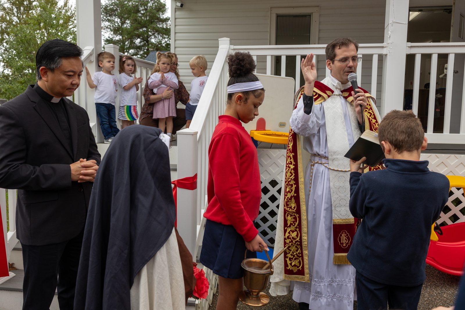 Father Scott Holmer, the pastor of Holy Face Parish in Great Mills, Maryland, blesses the new Early Learning Center at Little Flower School in Great Mills on Oct. 1, 2024, assisted by students Valencia Irving and Lawrence Pace. At left is Father John Nguyen, the pastor of St. George Parish in Valley Lee, Maryland, which cosponsors the school along with Holy Face Parish and Immaculate Heart of Mary Parish in Lexington Park. Standing on the porch in the background are four of the 2-year-old students who are attending the center. (Catholic Standard photo by Mihoko Owada)