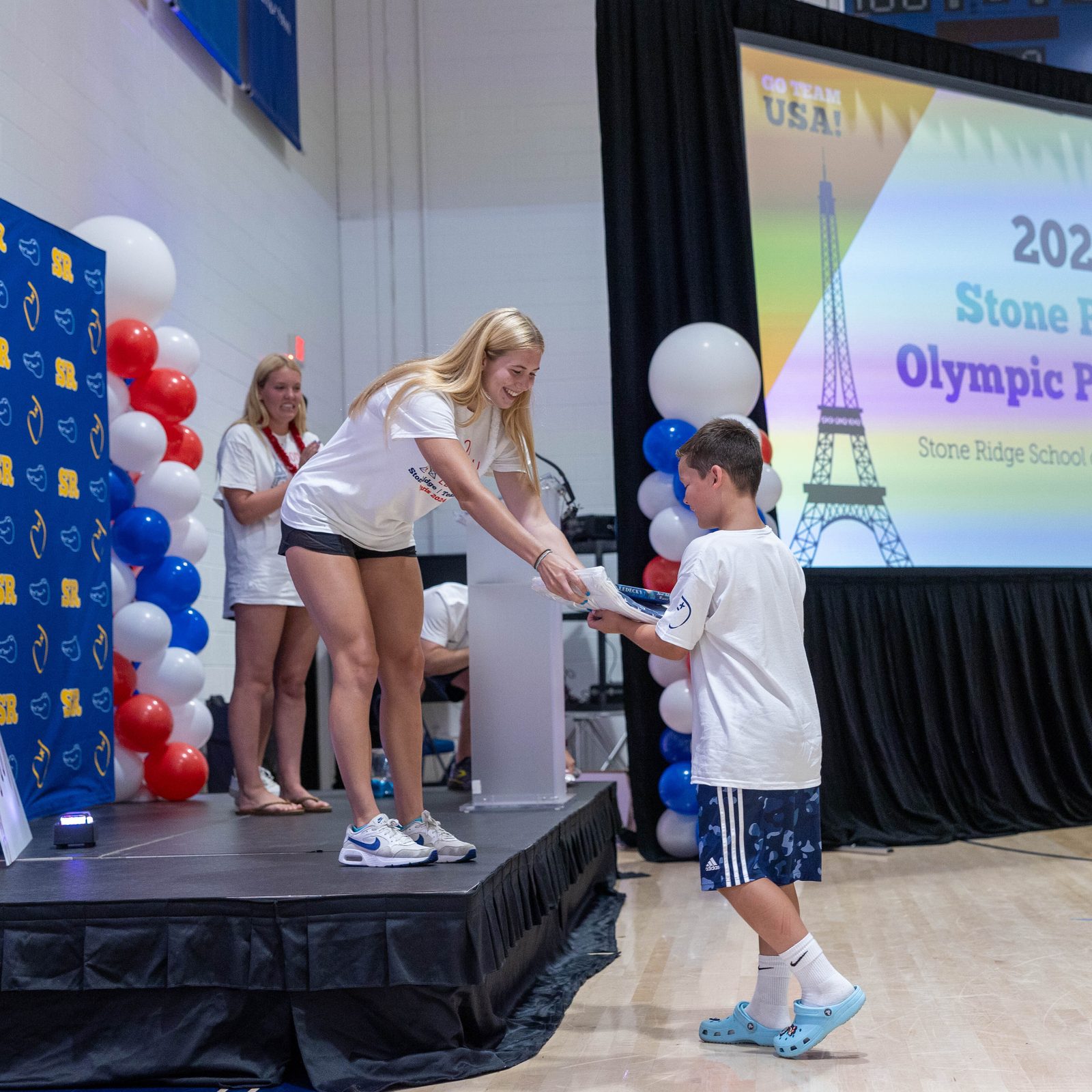 At a July 25 pep rally at Stone Ridge School of the Sacred Heart in Bethesda celebrating three of the school’s graduates who are competing in the 2024 Summer Olympics in Paris, Liza Goetcheus, a member of Stone Ridge’s class of 2023 who is currently a swimmer at Providence College, gives raffle prizes to a boy. The prizes included a recent autobiography written by Olympic swimming champion Katie Ledecky, a 2015 Stone Ridge graduate. The pep rally also celebrated Olympic swimmers and Stone Ridge graduates Phoebe Bacon (class of 2020) and Erin Gemmell (class of 2023). (Catholic Standard photo by Mihoko Owada)