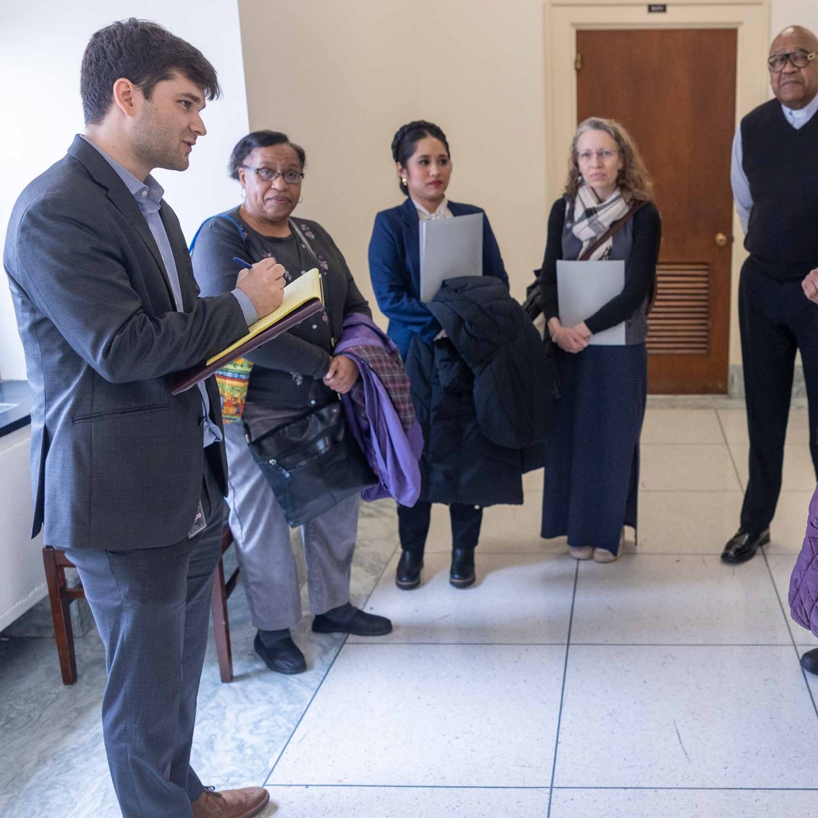 Sam Mollin, asistente legislativo del congresista Kweisi Mfume, (izq.), toma notas en la conversación con líderes religiosos y laicos de Baltimore que exponen su solidaridad con los inmigrantes, el 5 de marzo de 2026, en un pasillo del edificio congresional Rayburn en Capitol Hill. A la derecha se observa a la maestra Roxana Jara.