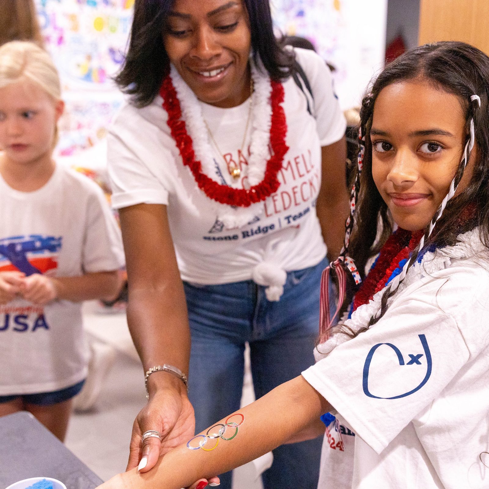 Before a July 25 pep rally at Stone Ridge School of the Sacred Heart in Bethesda celebrating three of the school’s graduates who are competing in the 2024 Summer Olympics in Paris, rising Stone Ridge fifth grader Madison Kunk, joined by her mother Sarah Kunk, shows the temporary tattoo on her arm displaying the Olympic rings symbol. Stone Ridge graduates  Katie Ledecky (class of 2015), Phoebe Bacon (class of 2020) and Erin Gemmell (class of 2023) are members of the U.S. Olympic Swimming Team competing in the Summer Olympics in Paris. In the photo below Lina Vuga (at left) a member of Stone Ridge’s class of 2024, helps summer camp student Viviana Bryant-Alcocer get ready for the pep rally. (Catholic Standard photos by Mihoko Owada)