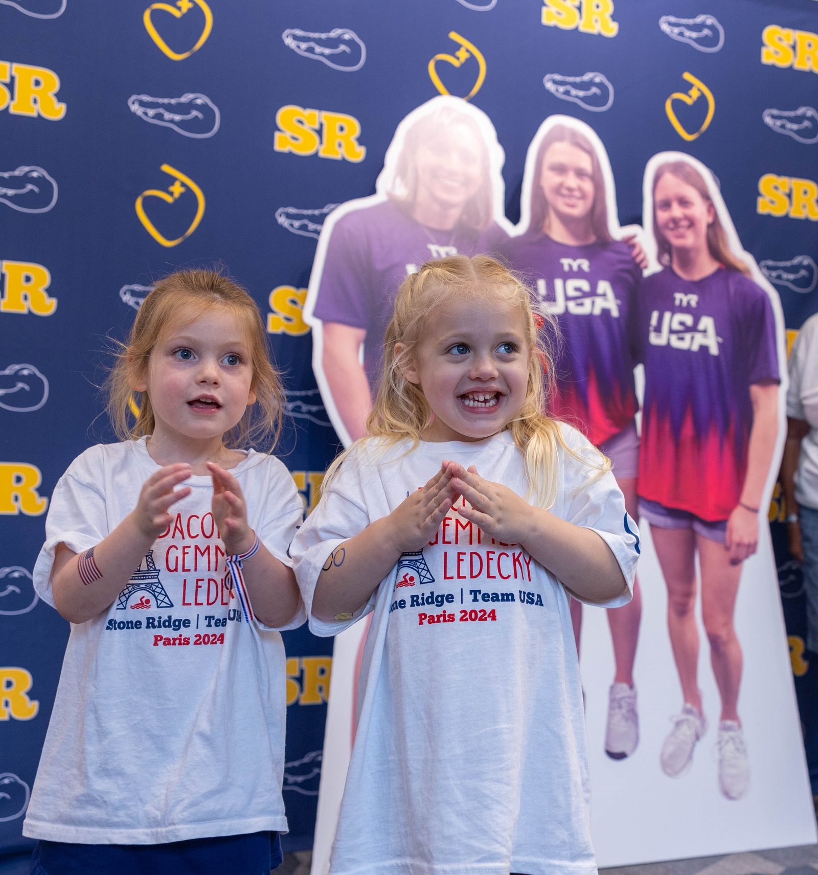 Stone Ridge pre-kindergarten students Clementine Ozinal, who is 4 years old, and Adeline Maguire, who is 3 years old, pose for a photo near the cutout images of the three graduates of their school who are swimming in the Paris Olympics. (Catholic Standard photo by Mihoko Owada)