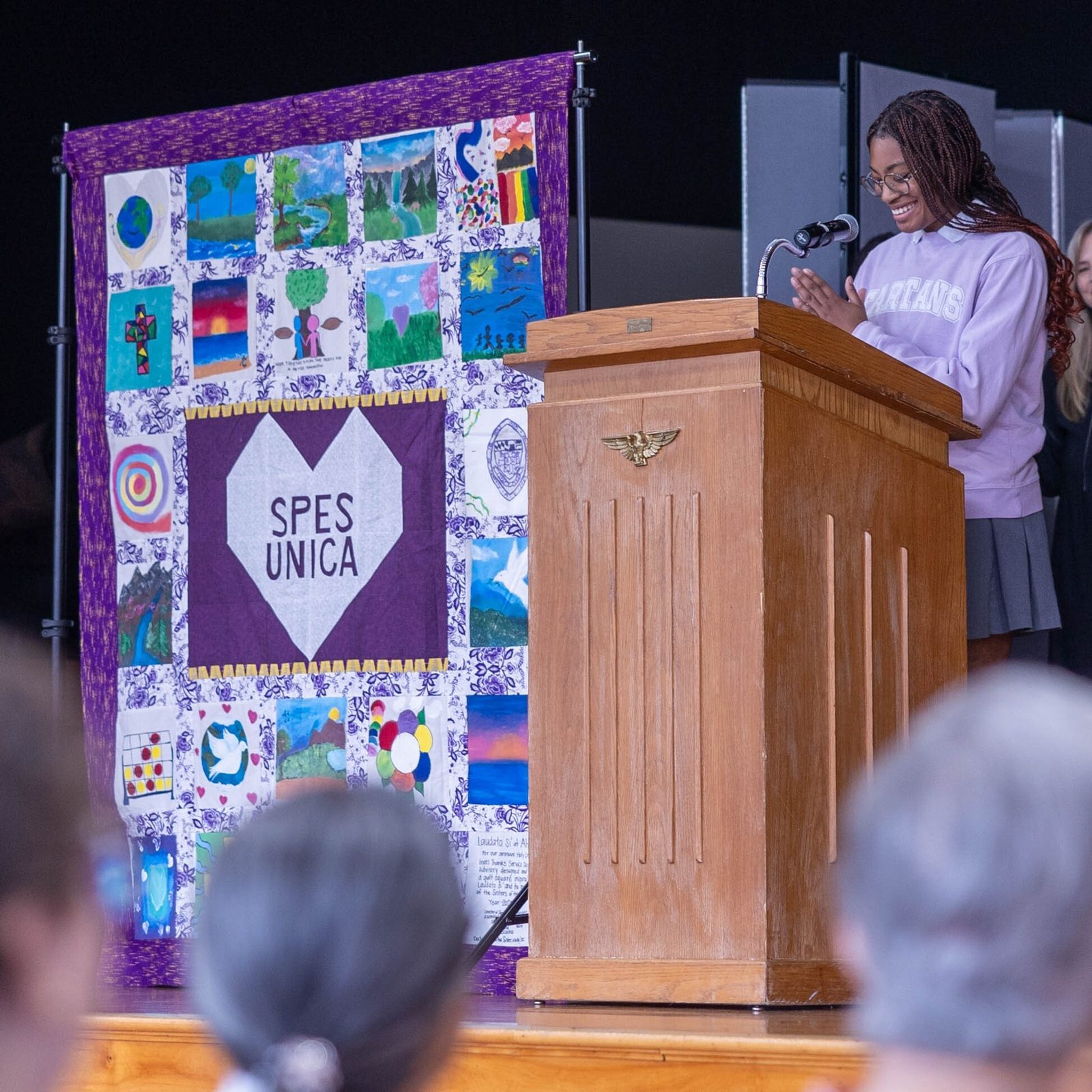 During a Sept. 13 Mass at The Academy of the Holy Cross in Kensington, Gabby Hill, a senior who serves as the Executive Board student president there, announces a quilt with hand-painted squares that students made for the Sisters of the Holy Cross to celebrate their work advancing peace and justice around the world. Standing at right is Kathy Hannah, the new principal at The Academy of the Holy Cross.  (Catholic Standard photo by Mihoko Owada)