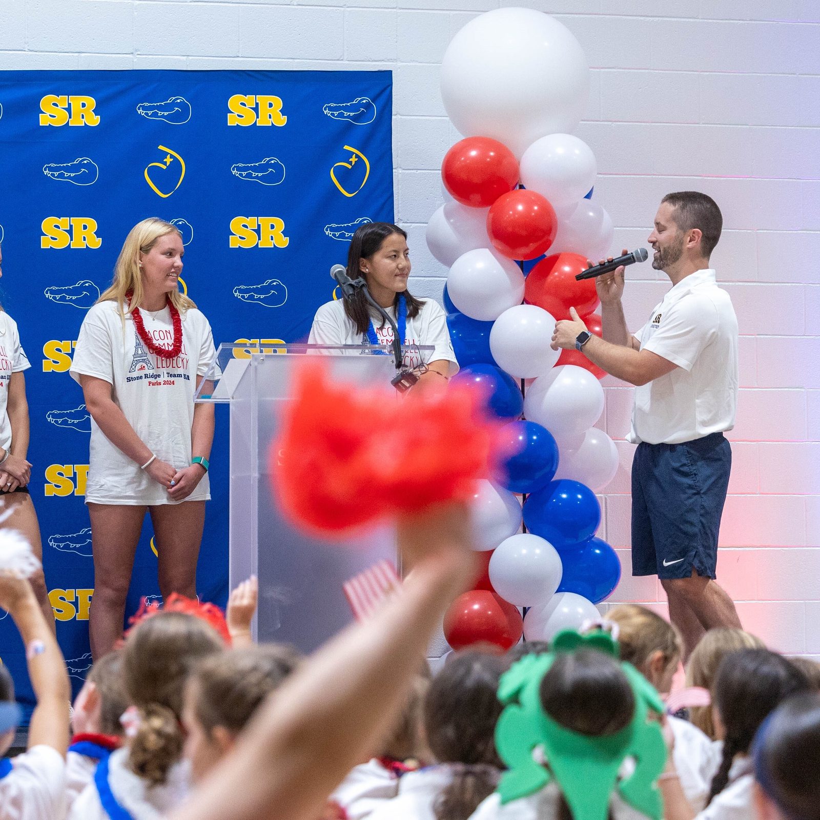 At a July 25 pep rally at Stone Ridge School of the Sacred Heart in Bethesda celebrating three of the school’s graduates who are competing in the Paris Olympics, Andrew Maguire (at right), the school’s director of athletics, introduces three Stone Ridge swimming team captains from the class of 2023 who spoke at the rally. From left to right are Liza Goetcheus, currently a swimmer at Providence College, Lauren Tucker, currently a swimmer at William & Mary, and Eleanor Sun, currently a swimmer at Princeton University. The pep rally celebrated Stone Ridge graduates Katie Ledecky (class of 2015), Phoebe Bacon (class of 2020) and Erin Gemmell (class of 2023), who are members of the U.S. Olympic Swimming Team competing in the Summer Olympics in Paris. (Catholic Standard photo by Mihoko Owada)